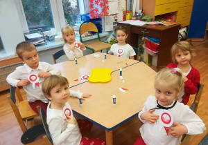 Szóstka dzieci przy stoliku prezentuje samodzielnie wykonane kokardy narodowe.